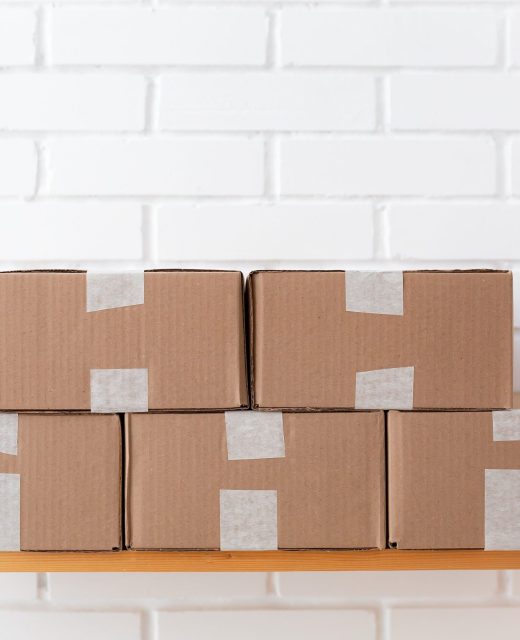 A neat stack of cardboard boxes taped and placed on a wooden shelf against a white brick wall.